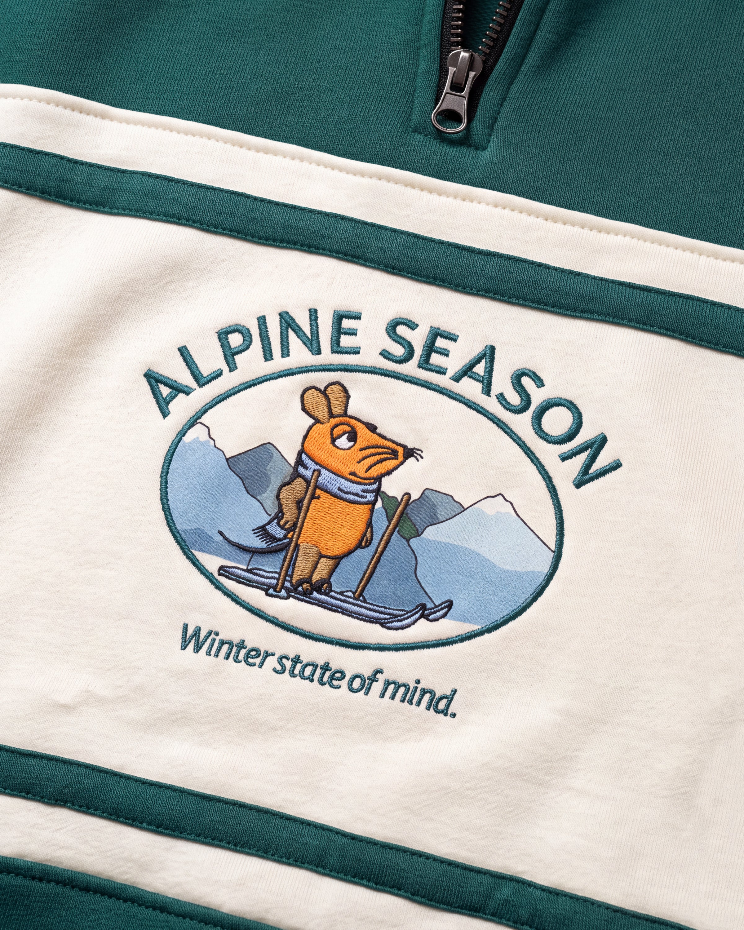 Detailaufnahme des Maus Alpine-Motivs auf dem grün-cremefarbenen Half Zip. Maus auf Skiern vor einer Bergkulisse.