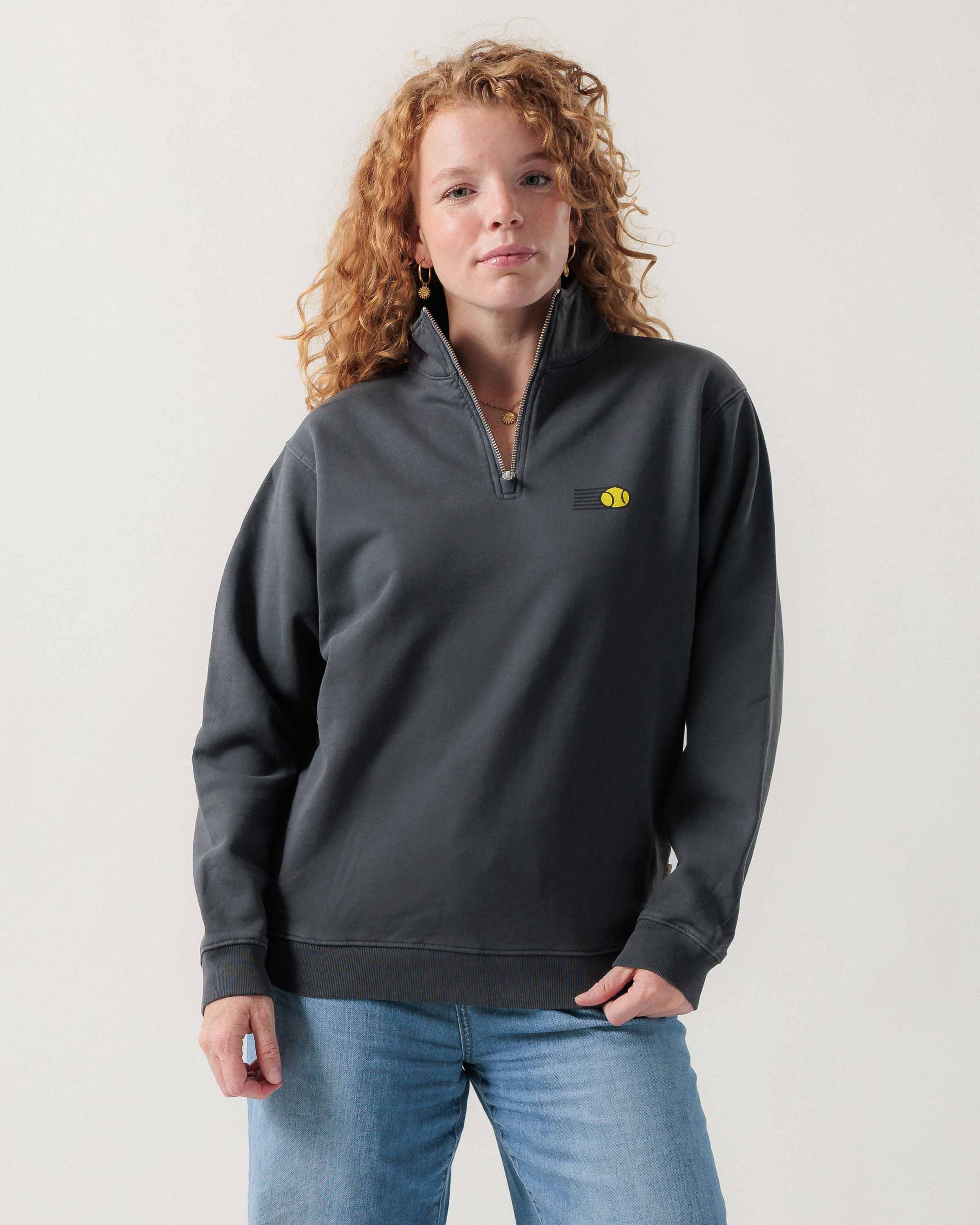Vorderansicht eines weiblichen Models im grauen Half Zip mit einem fliegenden Tennisball.