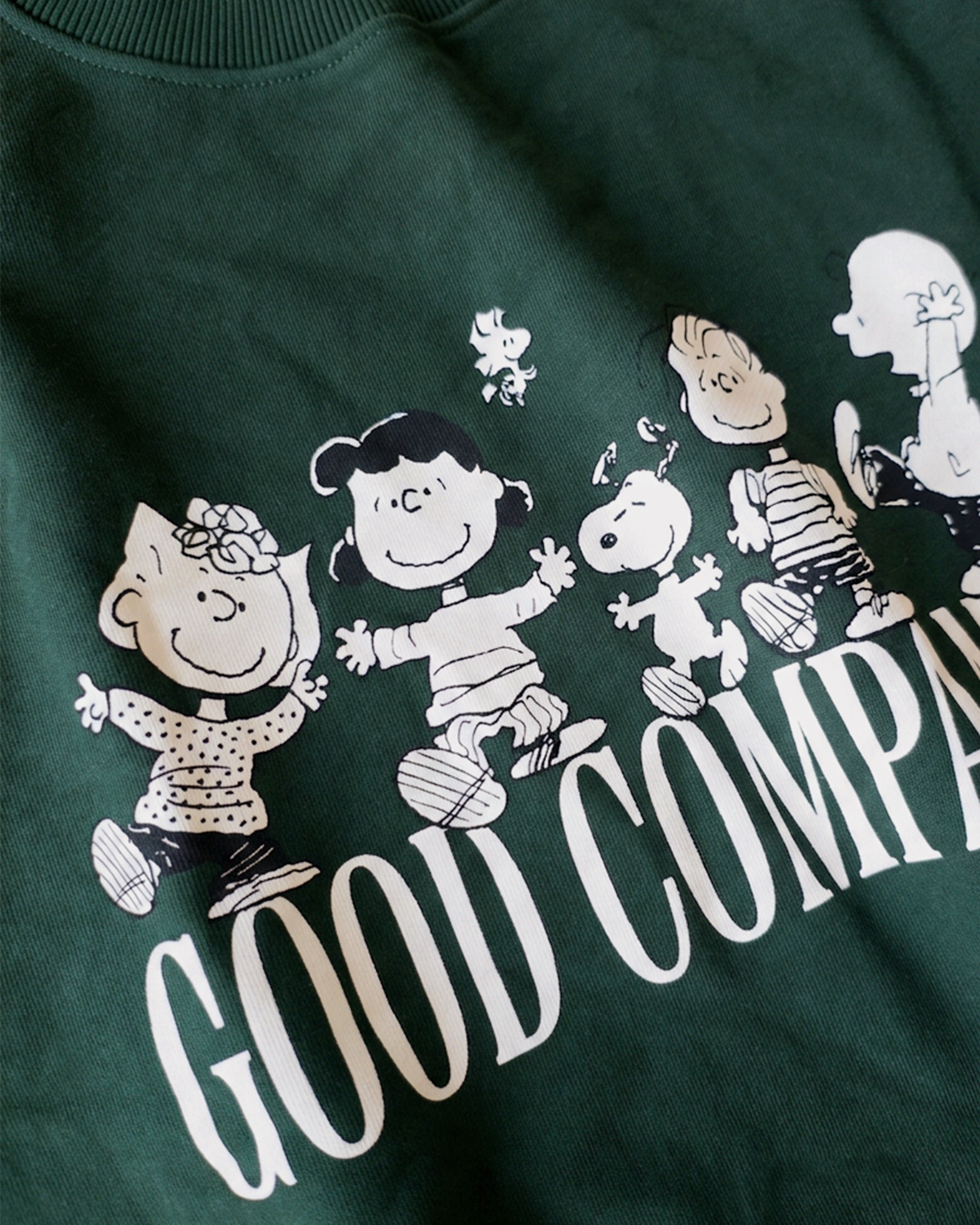 Detailaufnahme des Frontprints auf dem grünen Box Sweater. Snoopy und seine Freunde ausgelassen tanzend über dem „GOOD COMPANY“-Schriftzug.