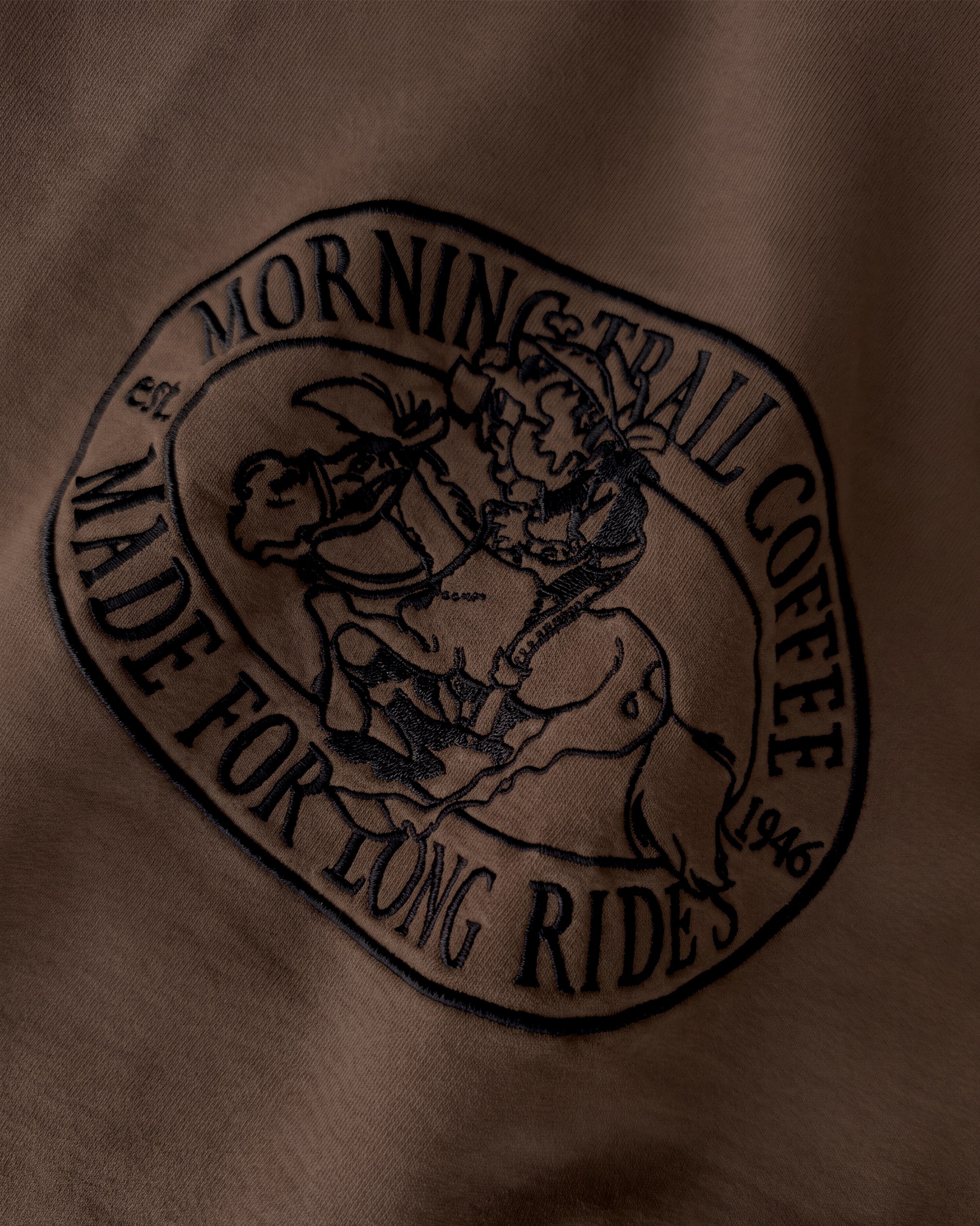 Detailaufnahme der Lucky Luke Coffee Run-Stickerei auf dem braunen Box Sweater. Lucky Luke, der auf Jolly Jumper reitend seinen Kaffee trinkt.