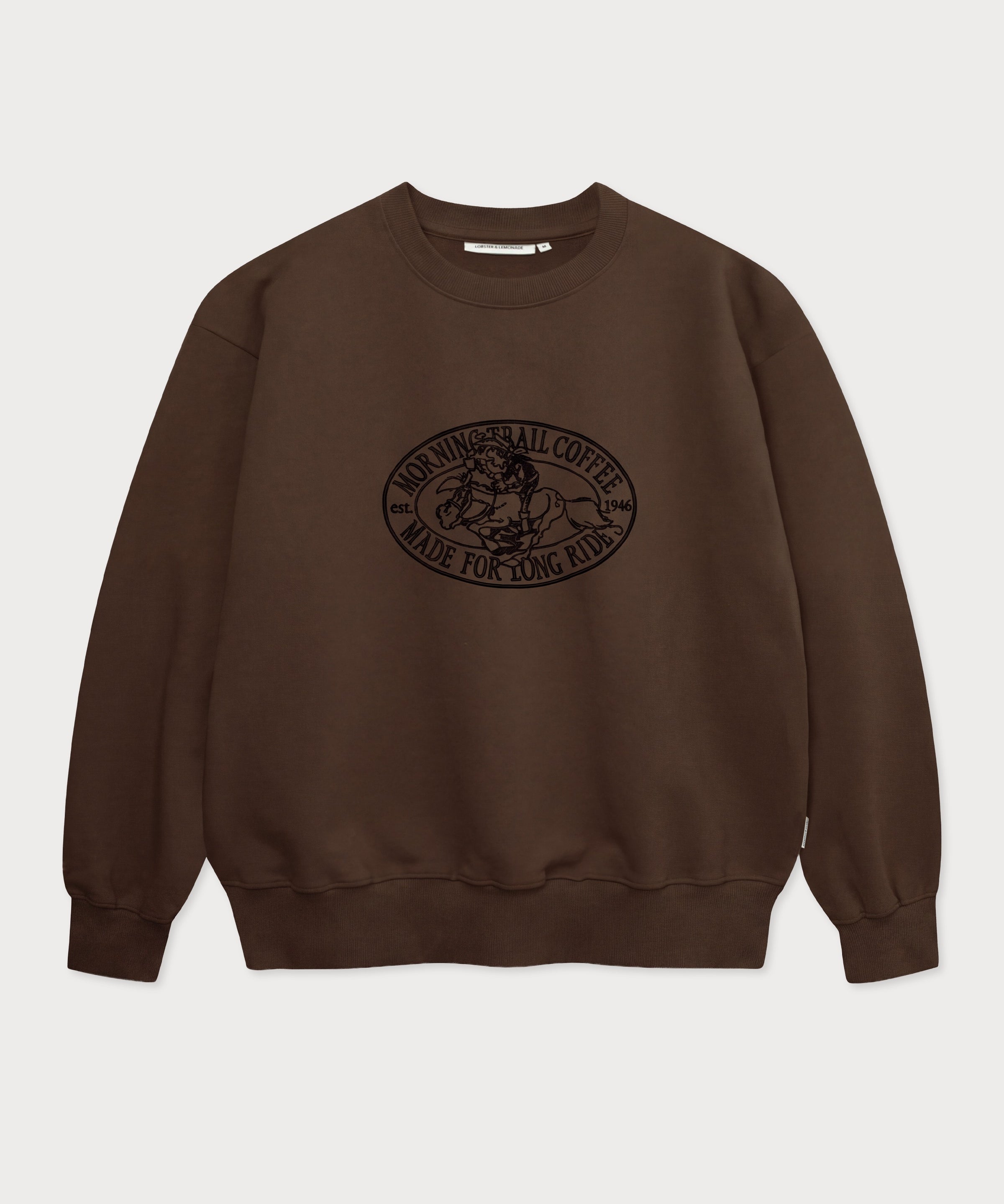 Vorderansicht des braunen Box Sweaters mit Lucky Luke Coffee Run-Stickerei. Lucky Luke, der auf Jolly Jumper reitend seinen Kaffee trinkt.