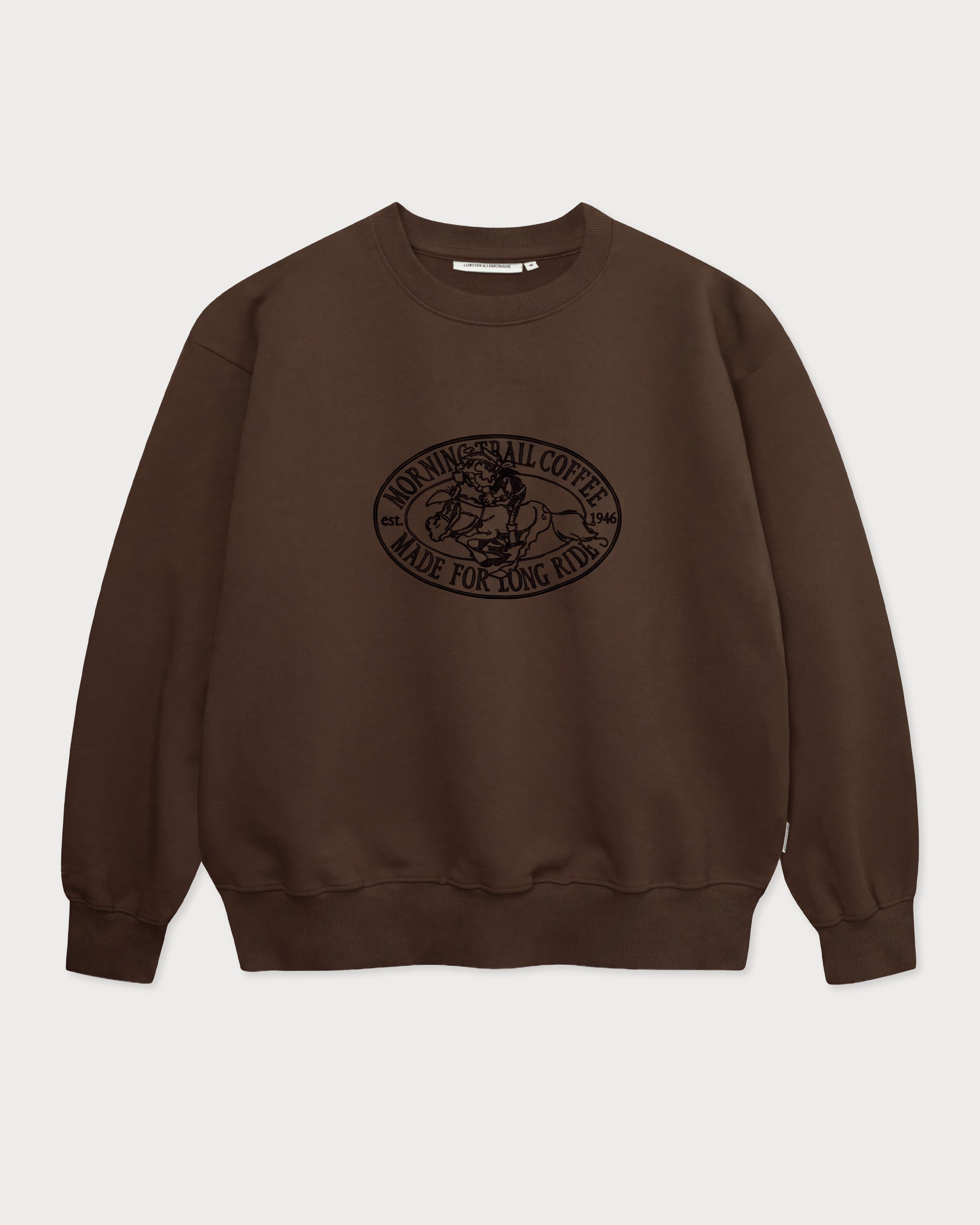 Vorderansicht des braunen Box Sweaters mit Lucky Luke Coffee Run-Stickerei. Lucky Luke, der auf Jolly Jumper reitend seinen Kaffee trinkt.