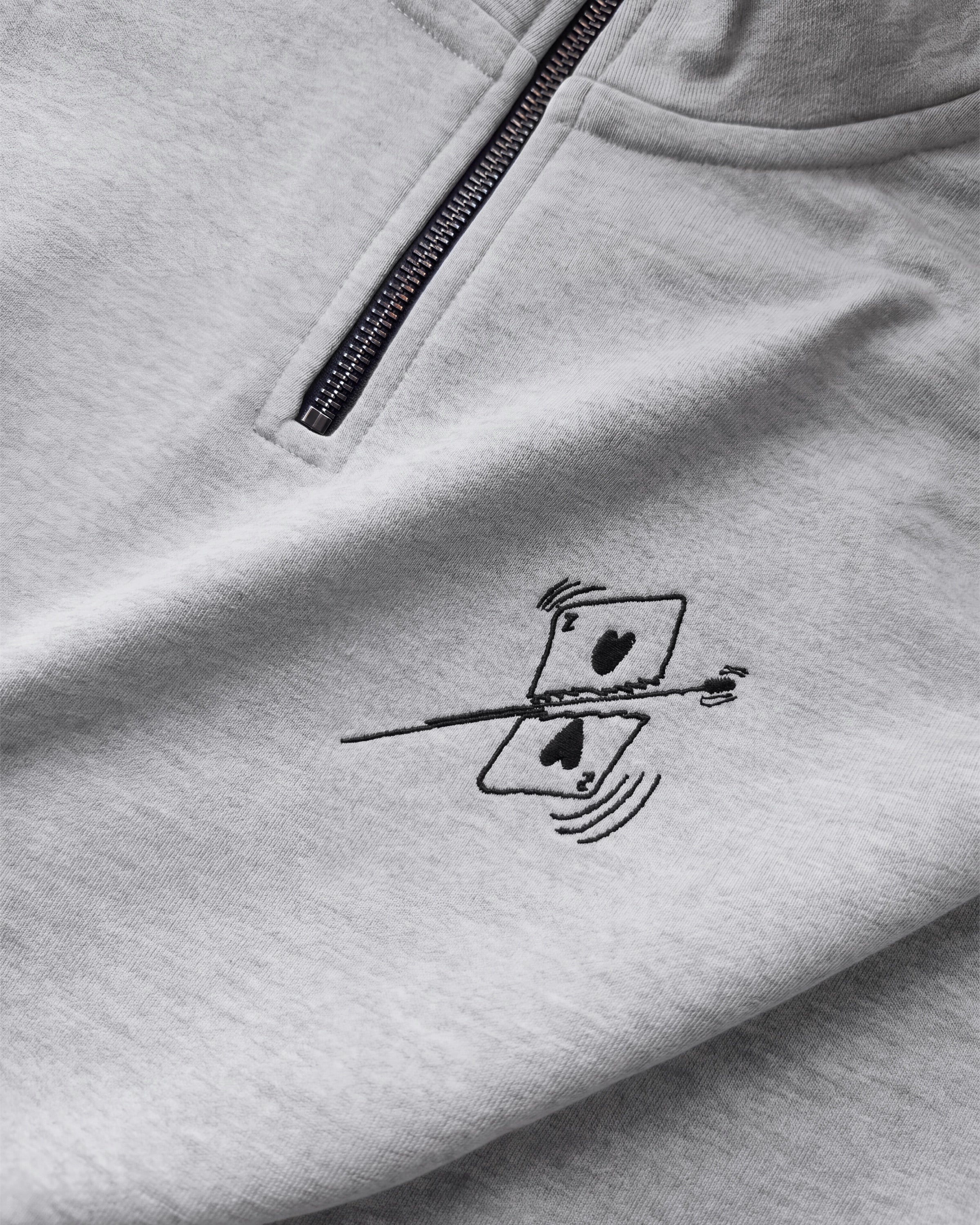 Detailaufnahme der Vorderseite des grauen Half Zips mit einer von einer Kugel durchbohrten Spielkarte auf der Brust.