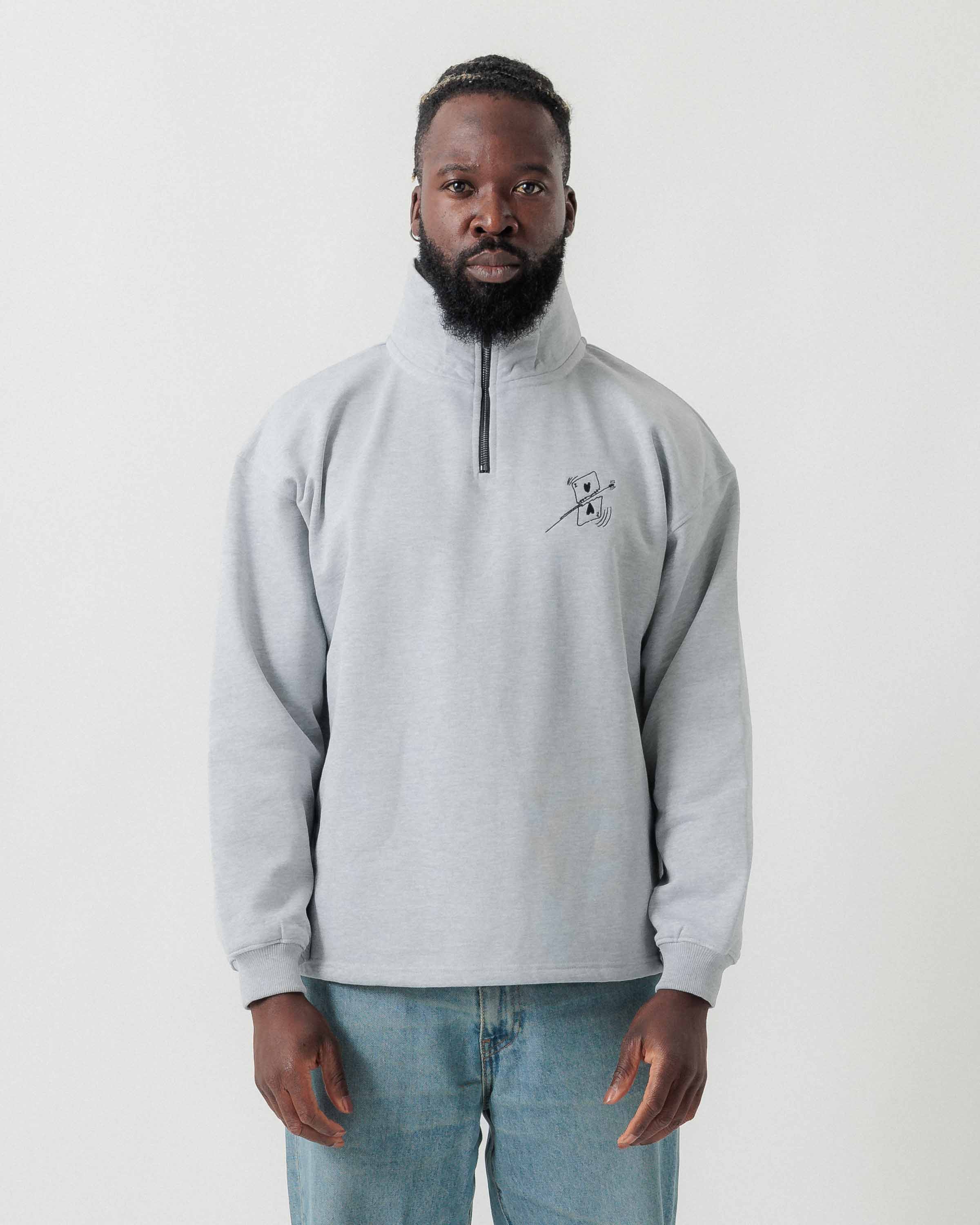 Vorderansicht eines männlichen Models im grauen Half Zip mit einer von einer Kugel durchbohrten Spielkarte auf der Brust.