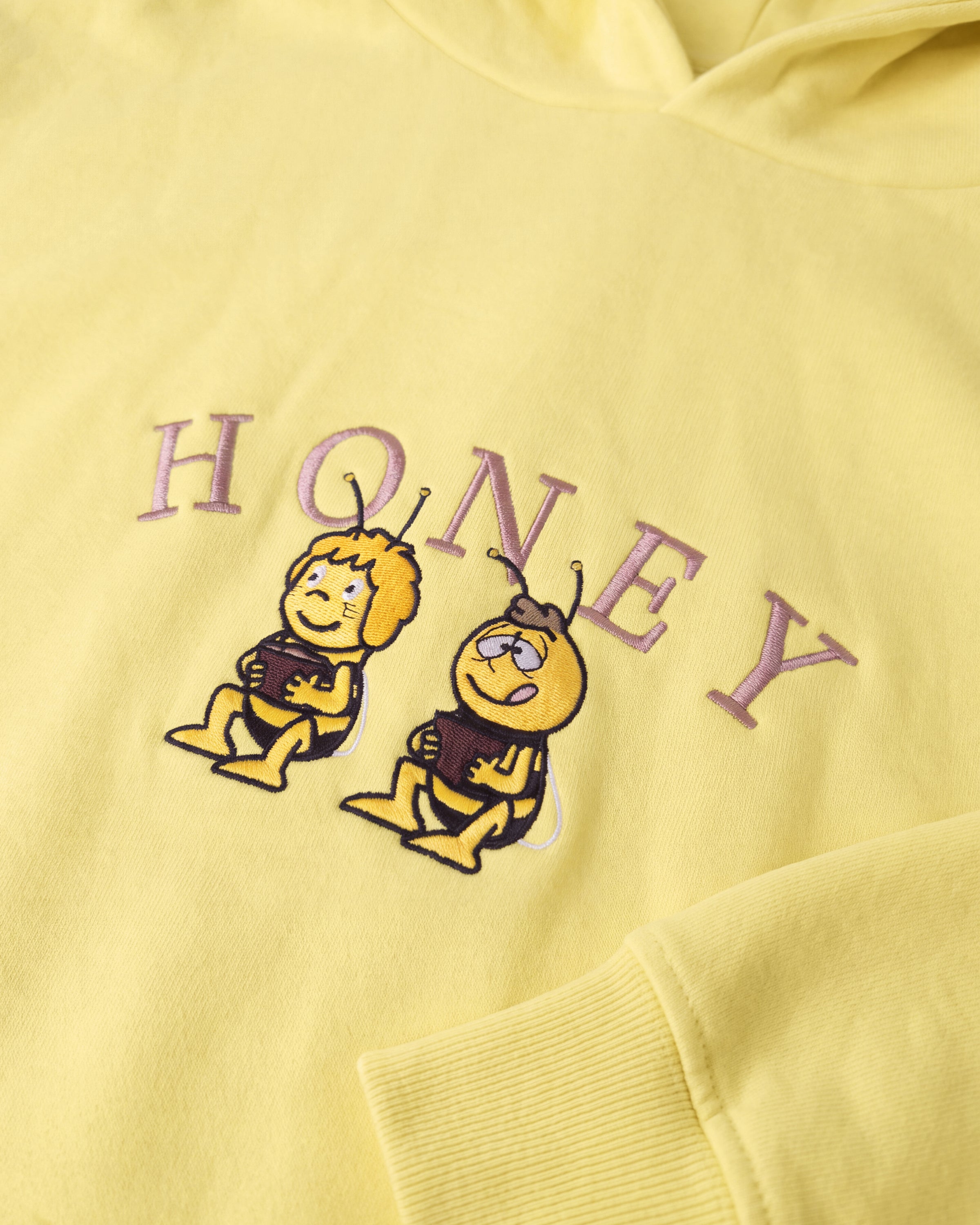 Detailaufnahme der Biene Maja Honey-Stickerei auf dem gelben Hoodie. Maja und Willi, die ihren Honig naschen.