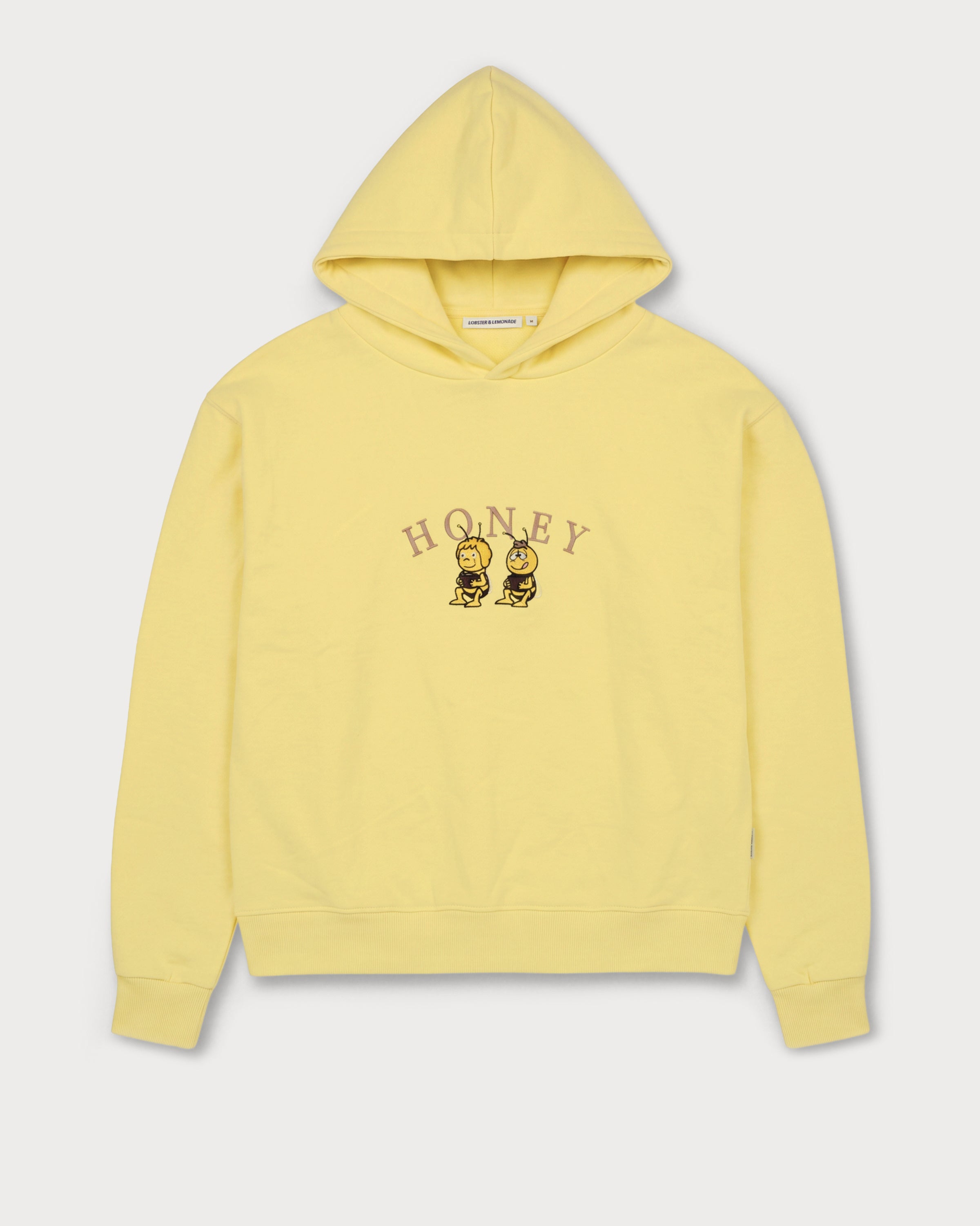 Vorderansicht des gelben Hoodies mit Biene Maja Honey-Stickerei auf der Brust. Maja und Willi, die ihren Honig naschen.