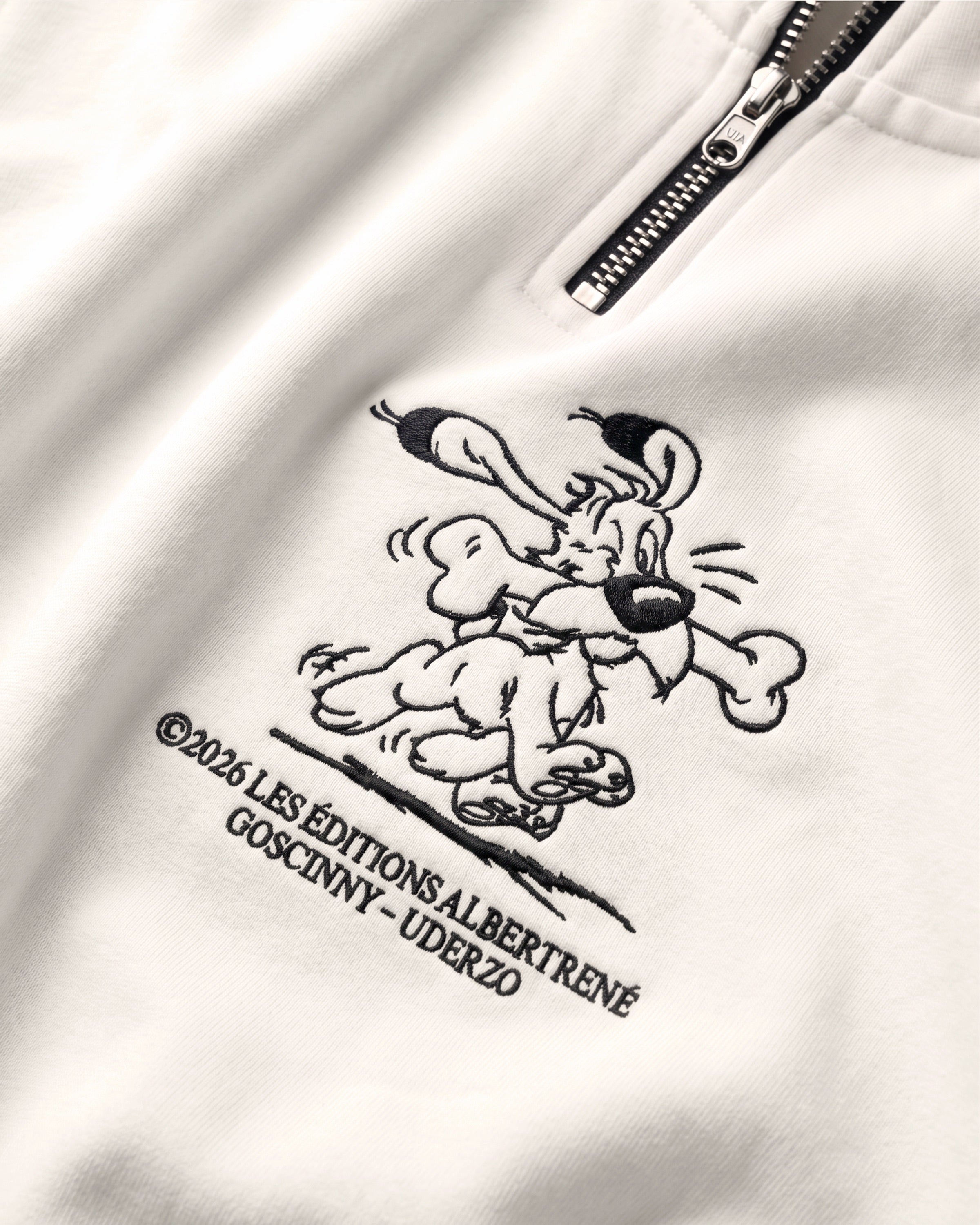 Detailaufnahme der Idéfix Happy-Stickerei auf dem weißen Half Zip. Der fröhliche Idefix mit einem Knochen.