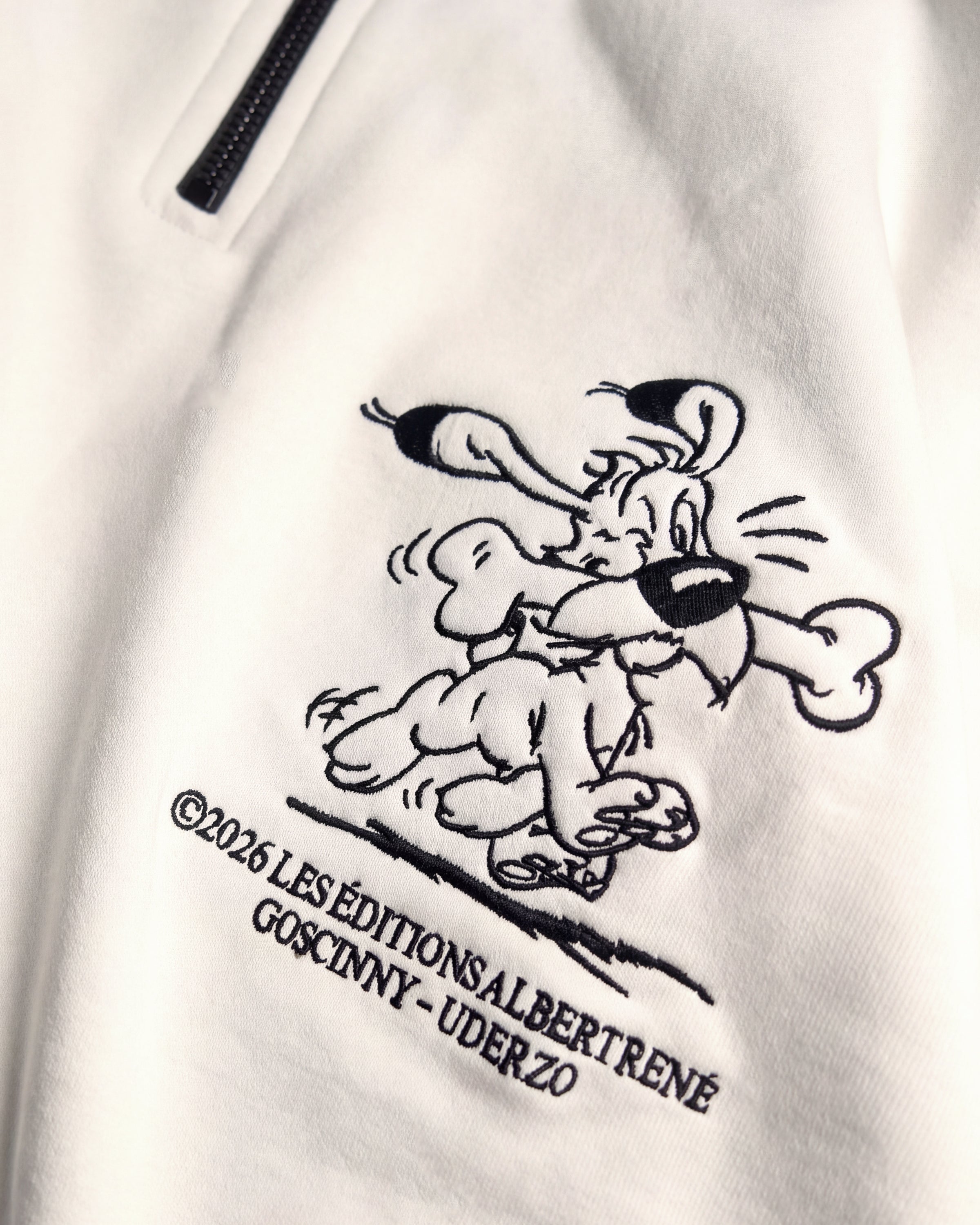 Detailaufnahme der Idéfix Happy-Stickerei auf dem weißen Half Zip. Der fröhliche Idefix mit einem Knochen.