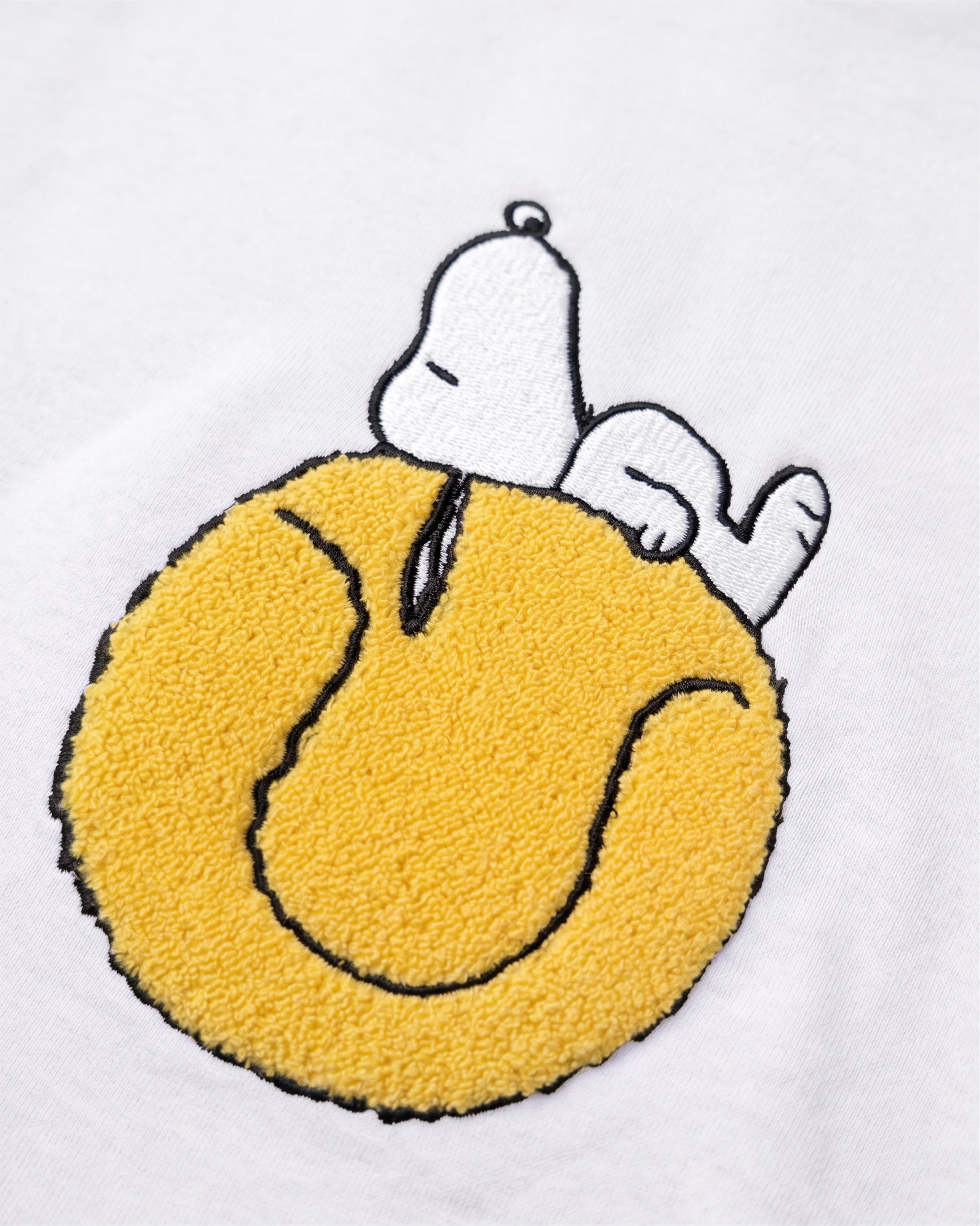 Detailaufnahme des Snoopy Tennisball-Motivs auf dem weißen Sweater. Tennisball, auf dem Snoopy tiefenentspannt relaxt.