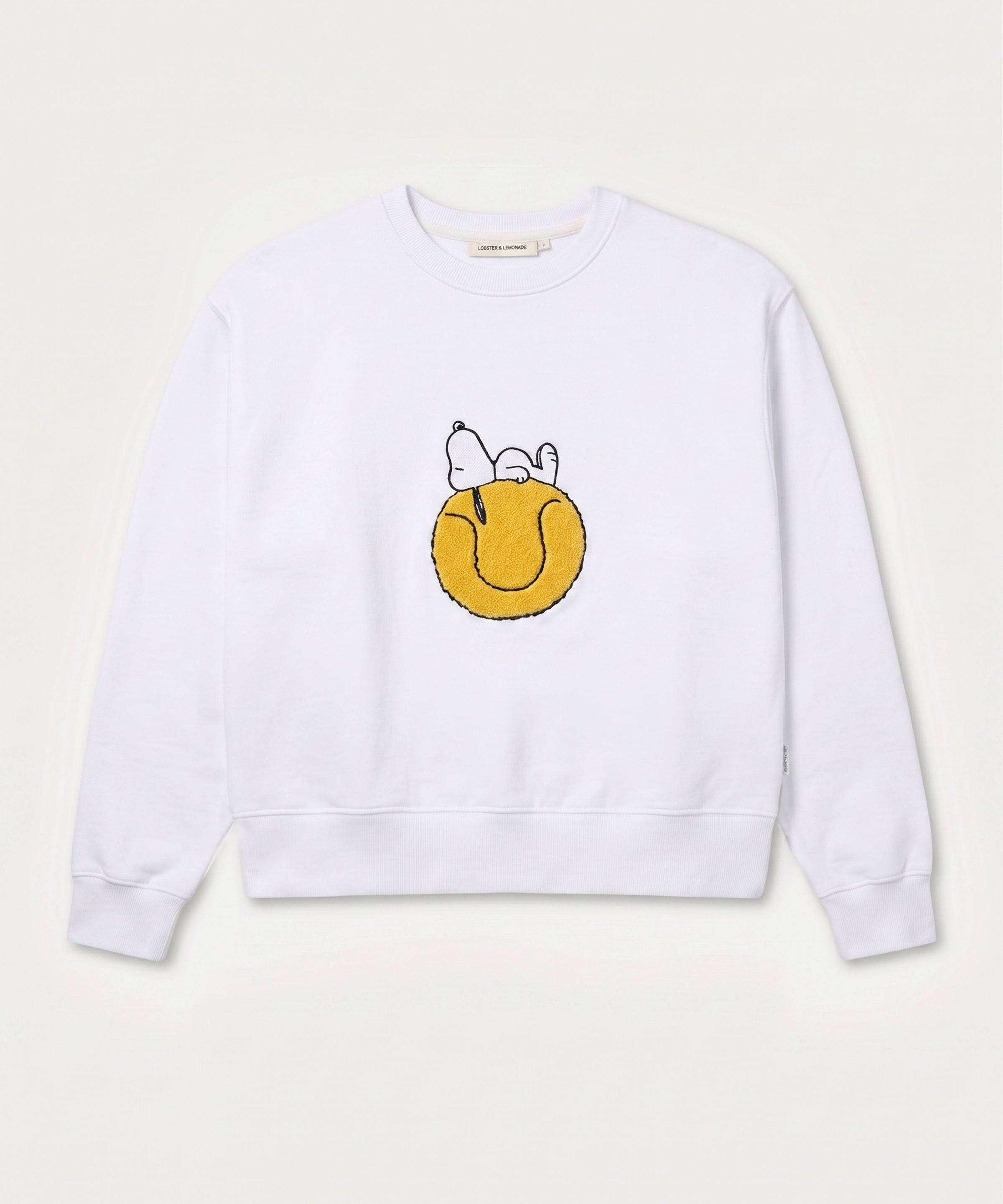 Vorderansicht des weißen Sweaters mit Snoopy Tennisball-Motiv. Tennisball, auf dem Snoopy tiefenentspannt relaxt.