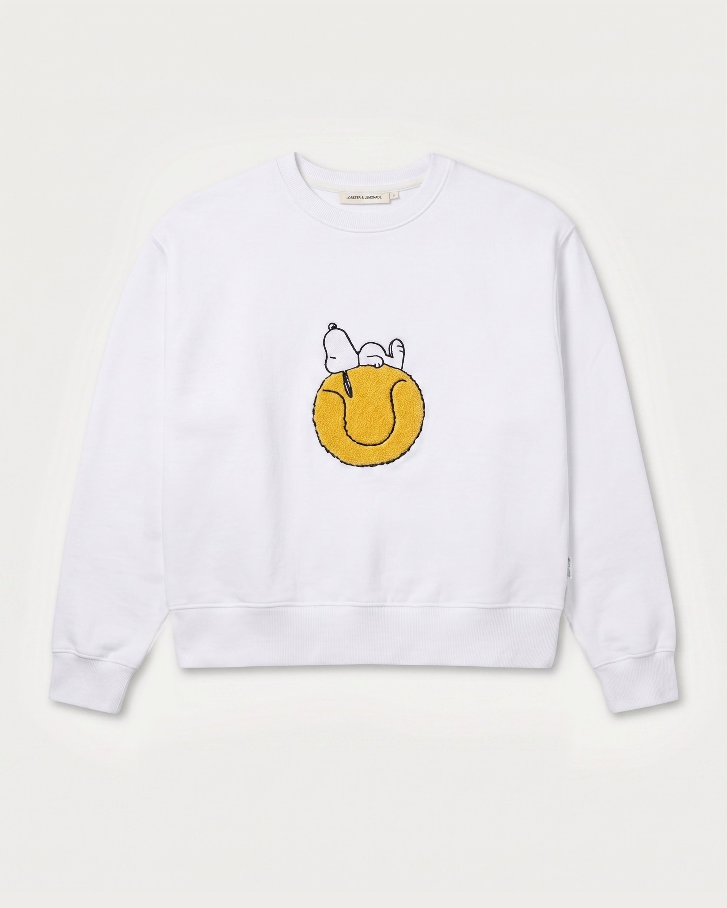 Vorderansicht des weißen Sweaters mit Snoopy Tennisball-Motiv. Tennisball, auf dem Snoopy tiefenentspannt relaxt.